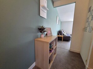 A hallway with a wooden console table, a potted plant, and a framed picture. There is a couch and a small table in the background. - Les toits de nationale, 2 personnes (Lille)