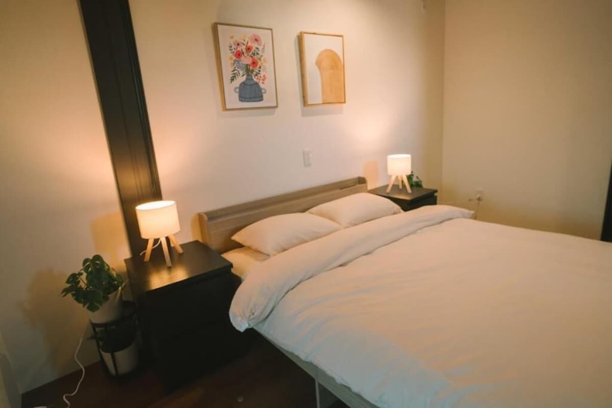 A bedroom with a bed, two bedside tables, a nightstand, a lamp, and a potted plant.
