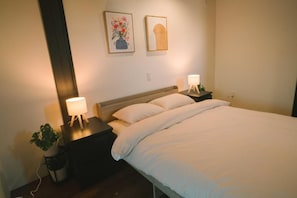 A bedroom with a bed, two bedside tables, a nightstand, a lamp, and a potted plant. - Seeds Stay Ijiri 2 (Fukuoka)