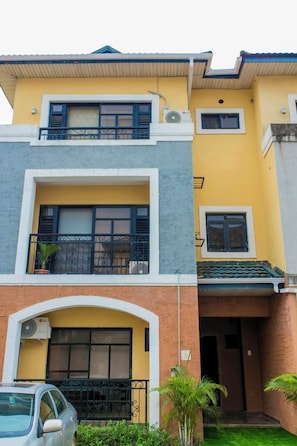Ein zweistöckiges Gebäude mit gelber Fassade, einem Balkon mit schwarzen Geländern und einem davor parkendem Auto.