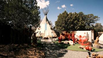 Un tipi tradicional con techo cónico, rodeado de un área boscosa con árboles y cielo despejado.