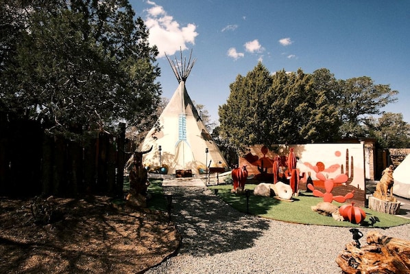 A traditional tipi with a conical roof, surrounded by a forested area with trees and a clear sky.