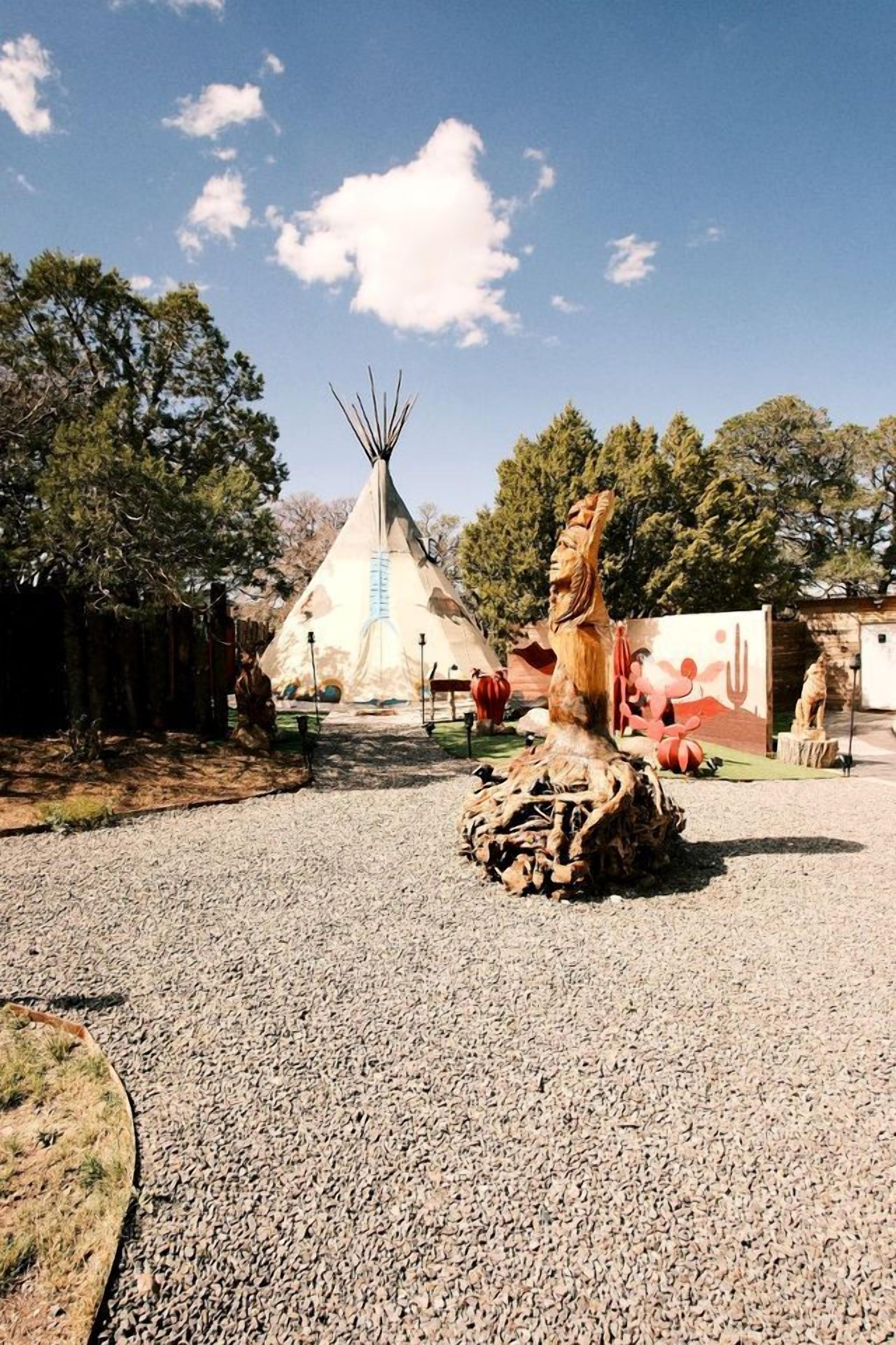 A traditional tipi with a blue stripe, surrounded by a gravel area with trees and a wooden fence.