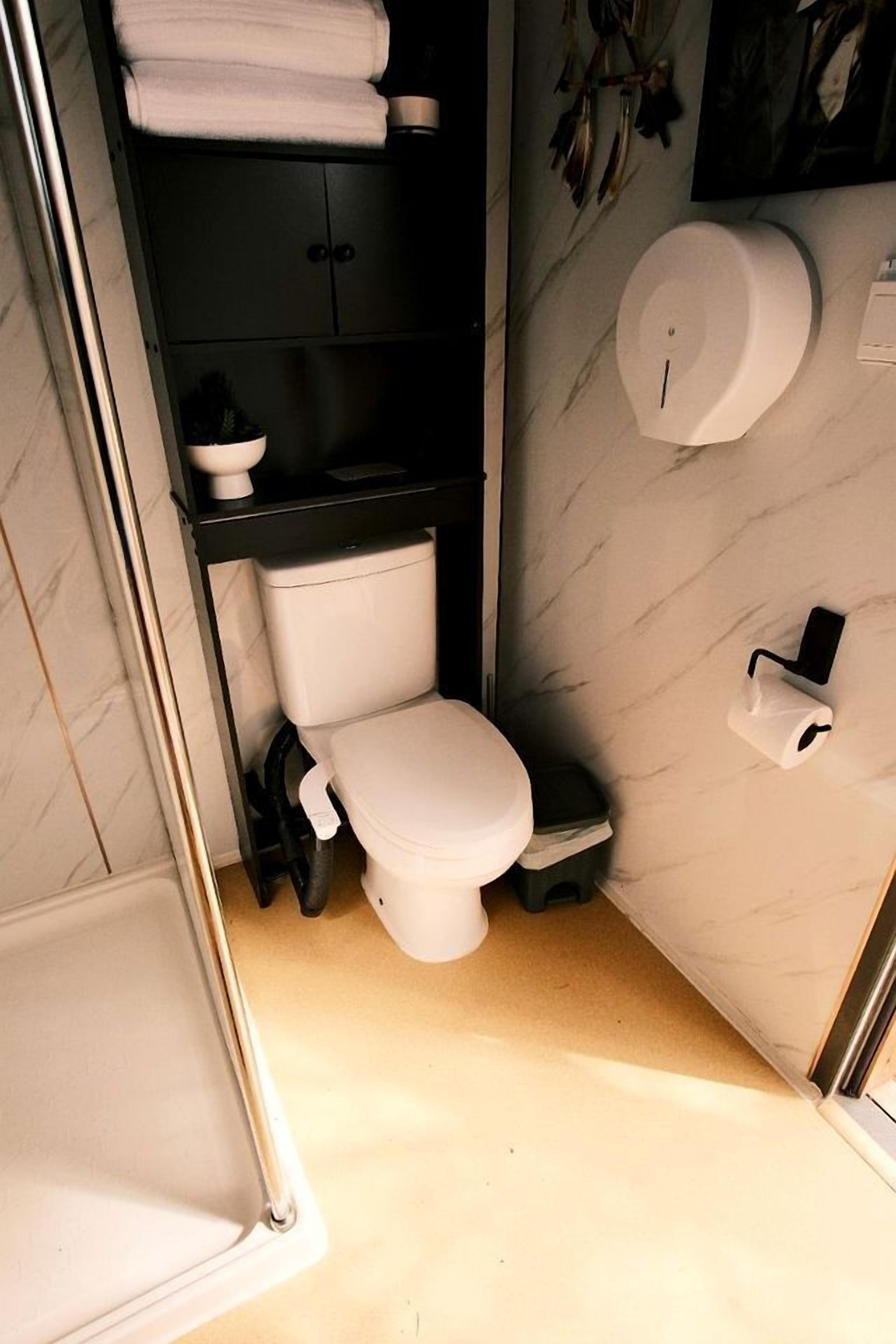 A compact bathroom with a toilet, a shelf with towels, and a wall-mounted toilet paper dispenser.