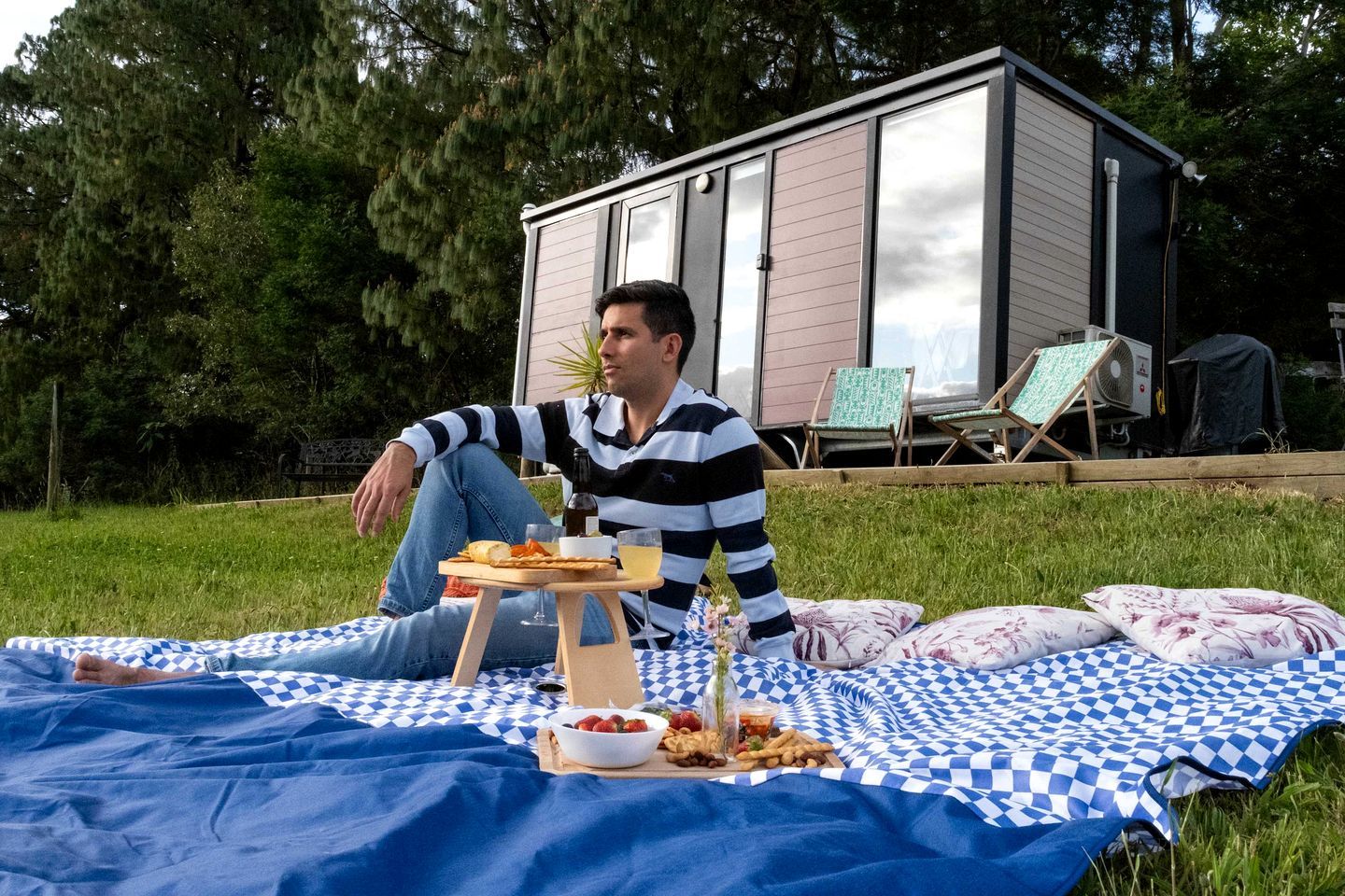 En person sidder på et ternet tæppe udendørs med et picnicbord, inklusive en flaske, glas og mad.