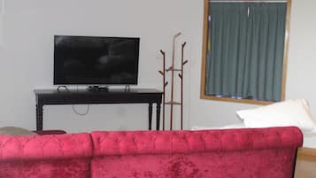 Una sala de estar con un sofá rojo, un televisor sobre un mueble bajo, una ventana con cortinas y un perchero.