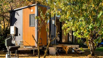 A compact, single-story mobile home with a wooden exterior and a small window, situated in a wooded area with trees and fallen leaves.