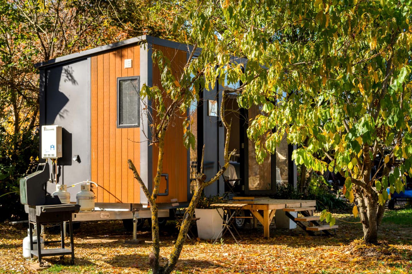 A compact, single-story mobile home with a wooden exterior and a small window, situated in a wooded area with trees and fallen leaves.
