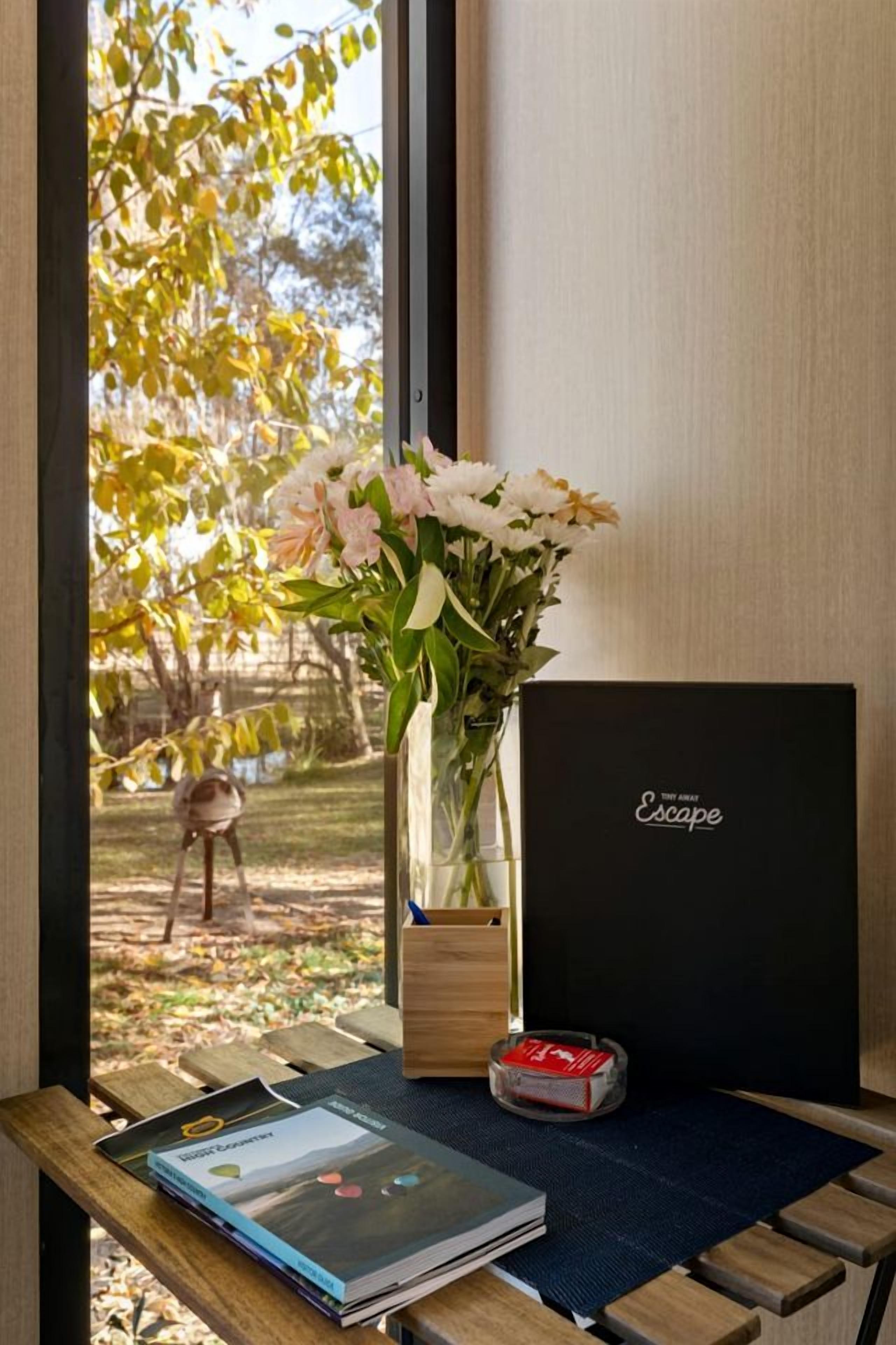A window with a view of trees, a vase with flowers, a book, and a black box on a wooden table.
