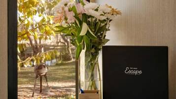 A window with a view of trees, a vase with flowers, a book, and a black box on a wooden table.