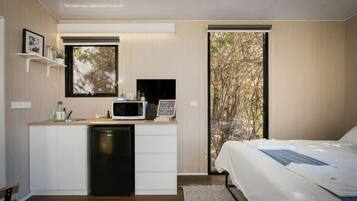 Ein modernes Schlafzimmer mit einem Bett, einem integrierten Schreibtisch mit Mikrowelle und Blick auf Bäume durch ein großes Fenster.