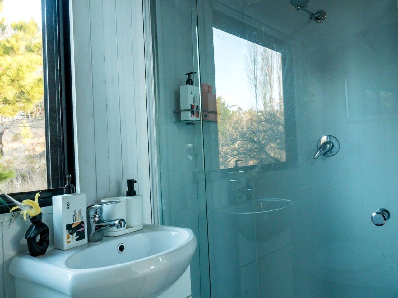 A modern bathroom with a glass shower enclosure, a white sink, and a window providing a view of trees.