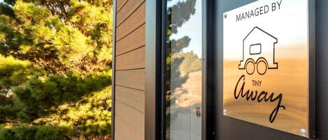 A modern building with a metal sign that reads 'MANAGED BY TINY Away' and a reflection of trees in the glass door.