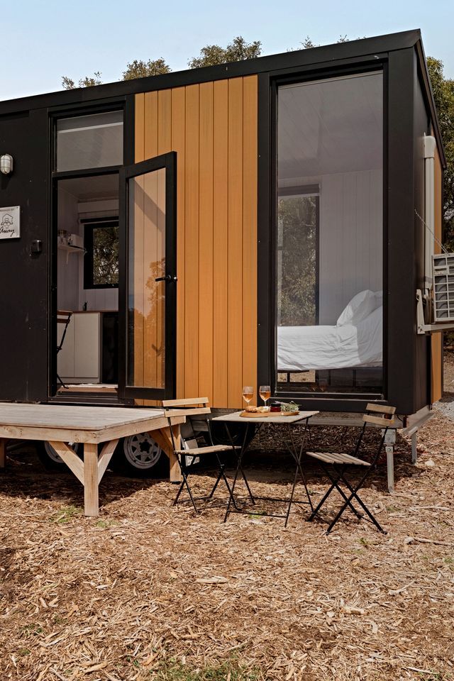 A small, modern cabin with a sliding door, a bed visible through the window, and a picnic table outside.