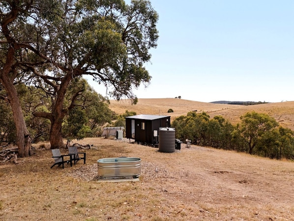 A rustic cabin with a water tank and a bench in a dry, hilly landscape.