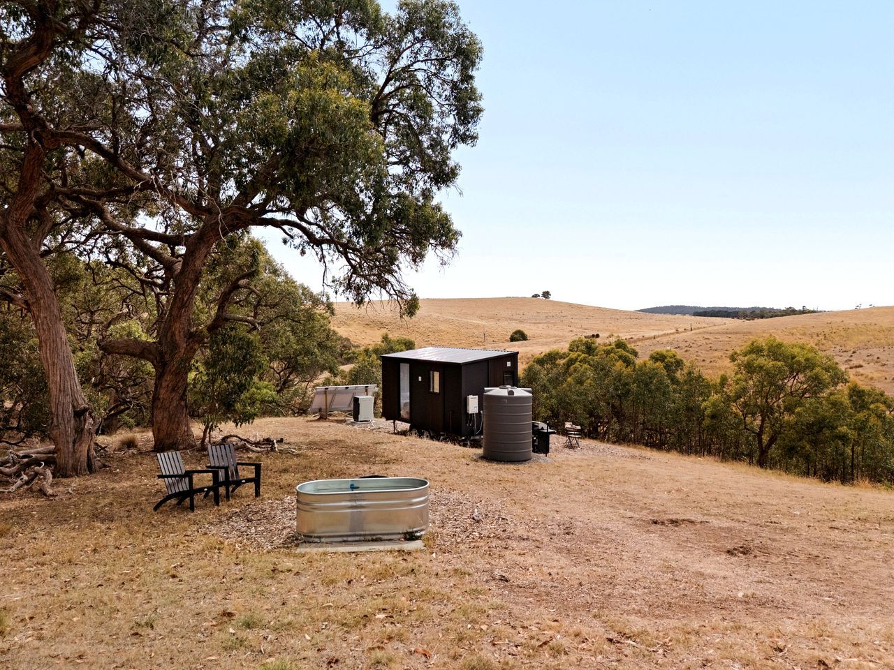 A rustic cabin with a water tank and a bench in a dry, hilly landscape.