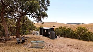 A rustic cabin with a water tank and a bench in a dry, hilly landscape.