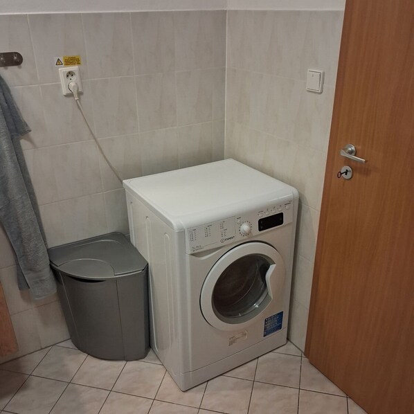 A compact, modern laundry room with a front-loading washing machine, a grey trash bin, and a door with a key lock.
