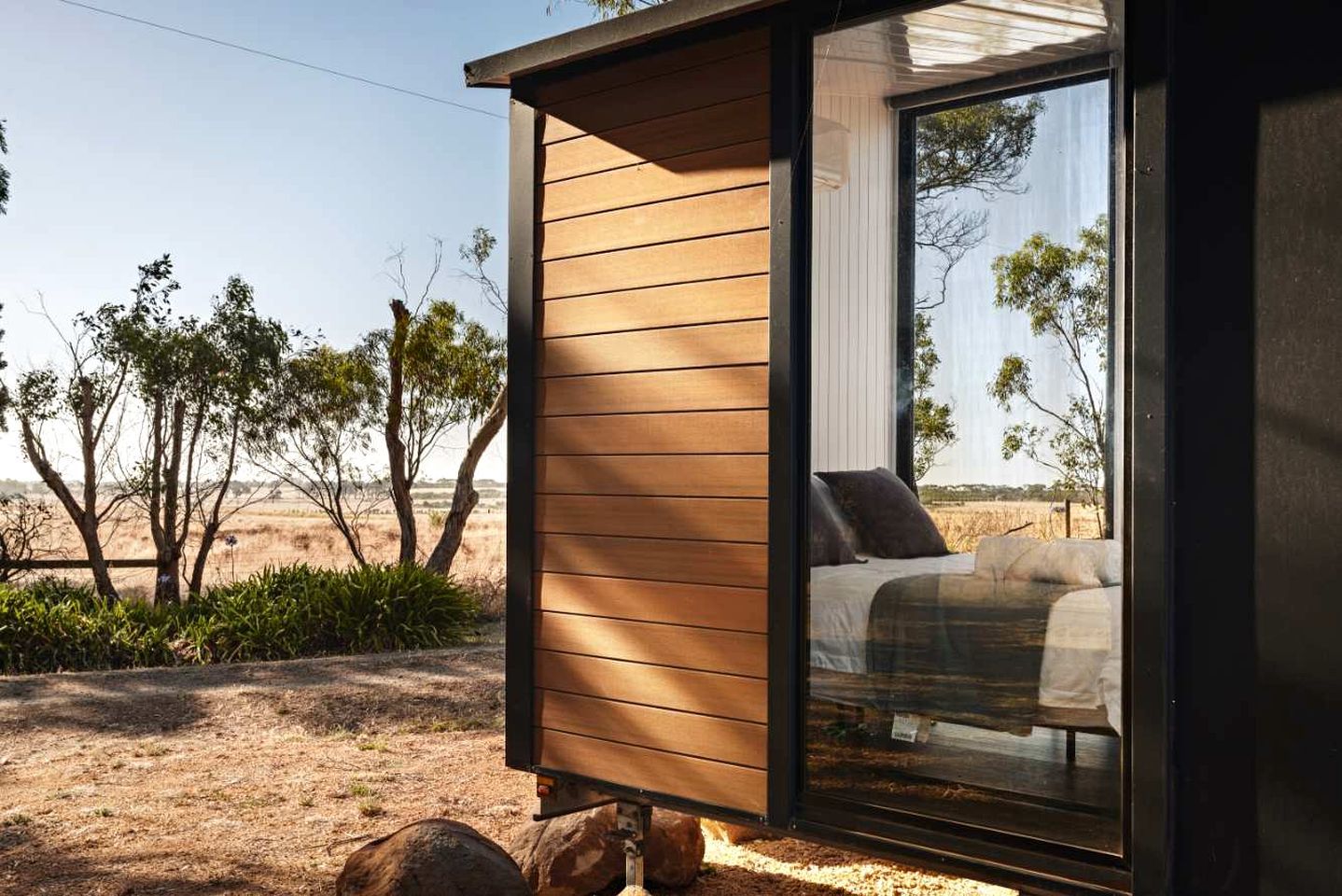 Ein modernes Schlafzimmer mit einem großen Schiebeglasfenster, einem Bett mit Kissen und Blick auf die umliegende Landschaft.