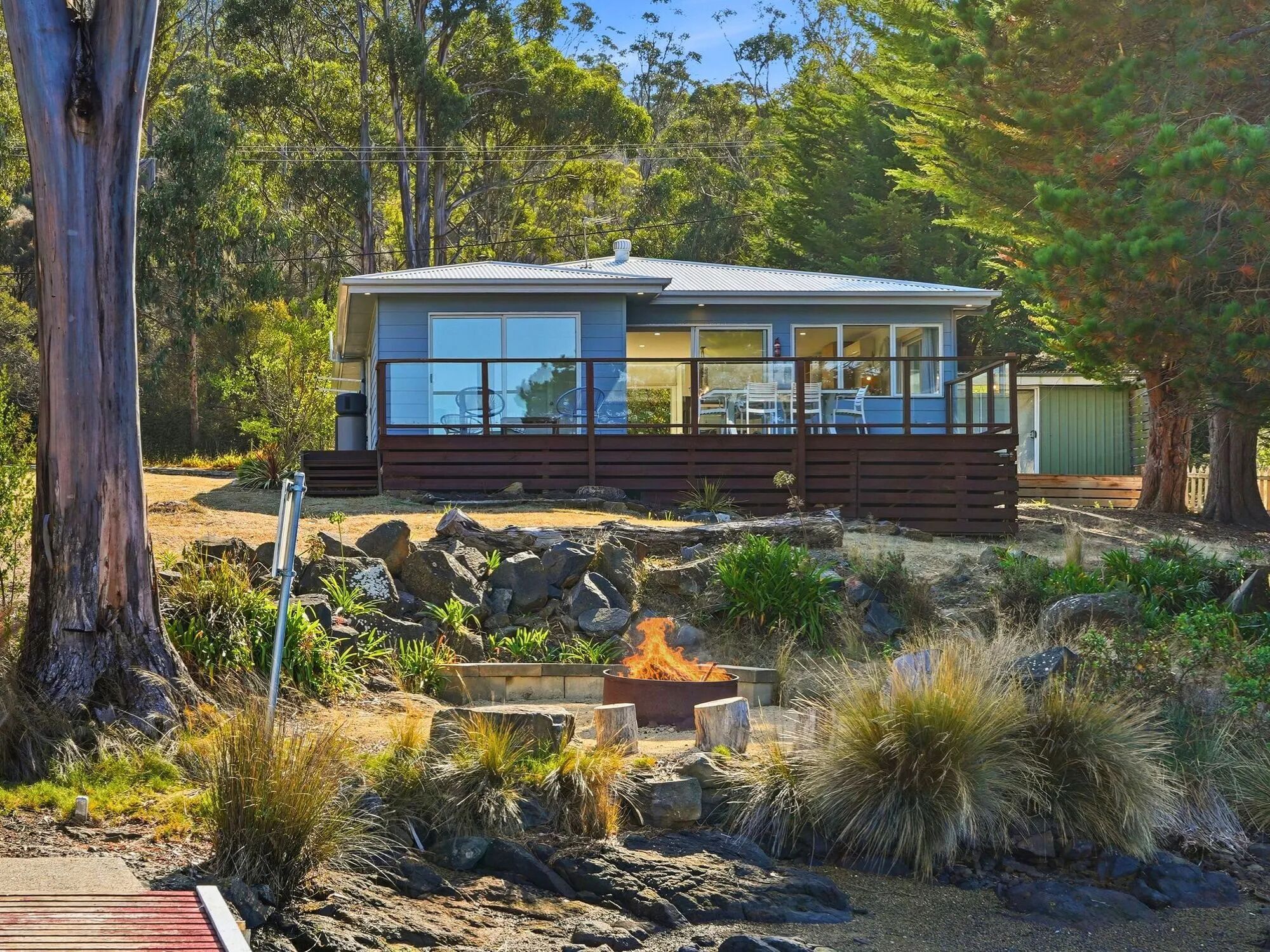 Une maison moderne dotée d’une terrasse, entourée d’une végétation luxuriante et agrémentée d’un foyer extérieur.