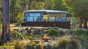 A modern house with a deck, surrounded by natural vegetation and a fire pit.