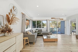 A spacious living room with a grey sectional sofa, a coffee table, and a large window providing a view of the outdoors. - Prosser River Retreat - Gateway to Maria Island (Orford)