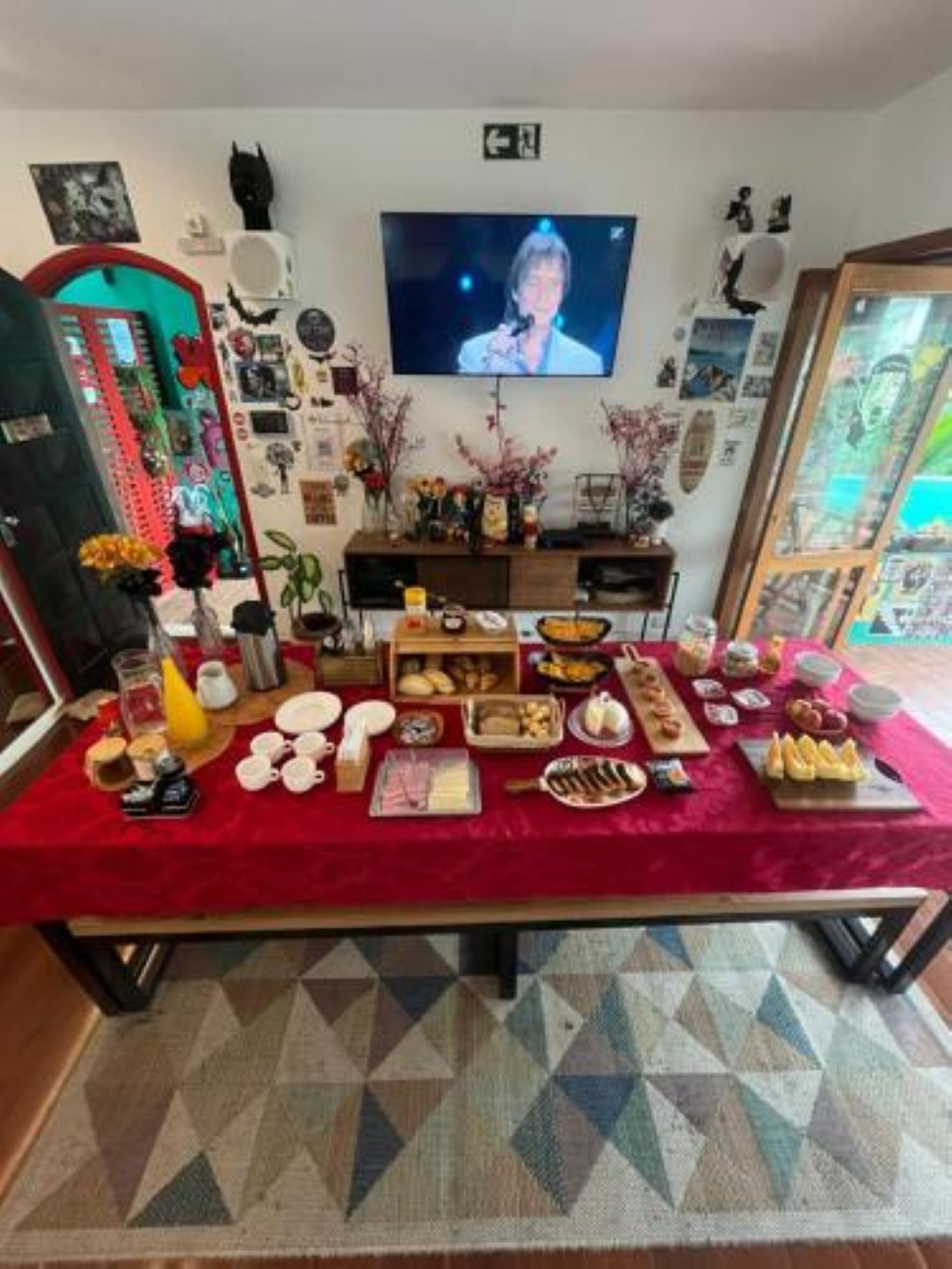 A room with a television mounted on the wall, a table with various food items, and a patterned rug.