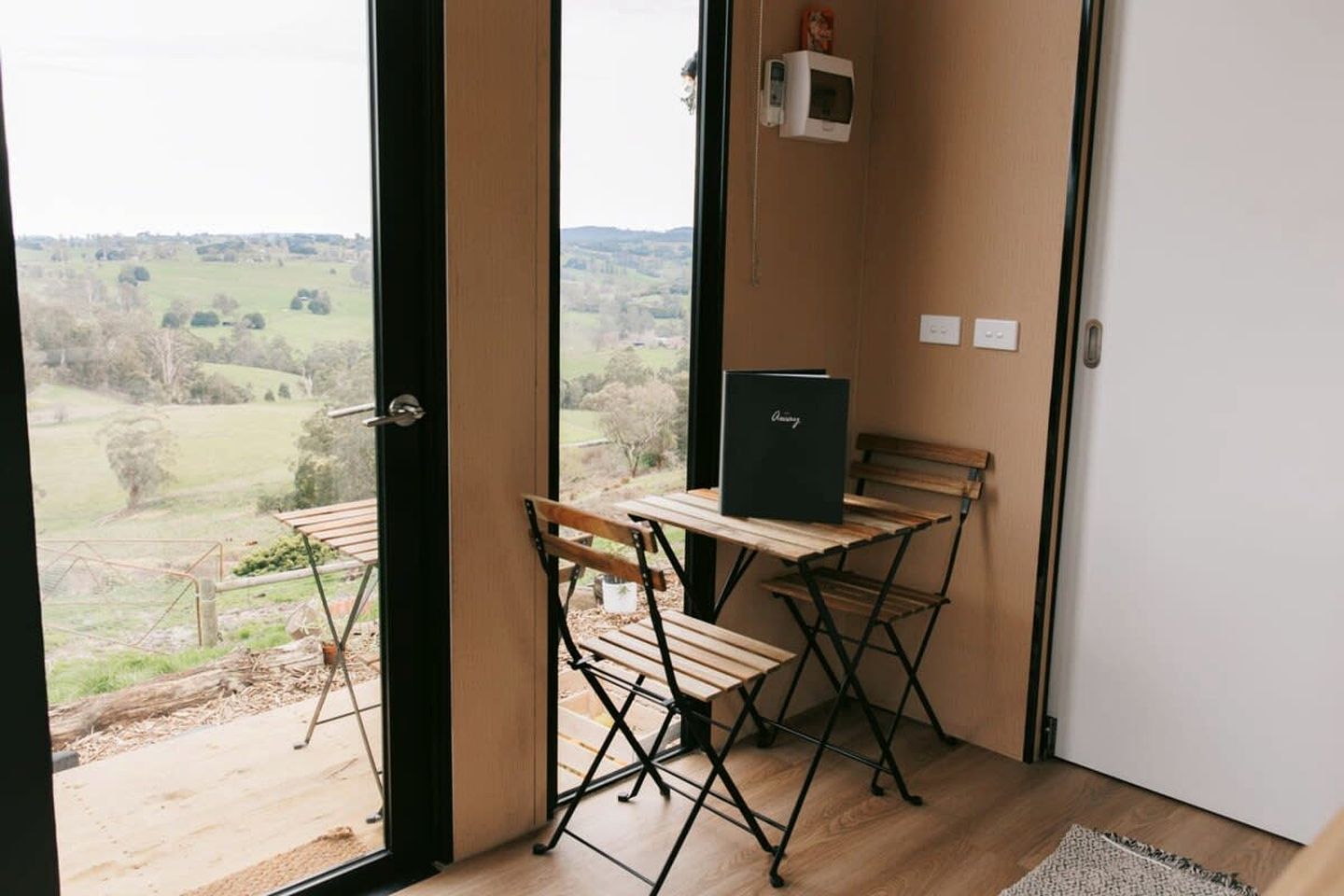 A room with a view of a rural landscape, a wooden table with a black folder, and a bench.