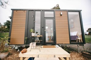 A modern, compact mobile home with a large sliding door, a small outdoor seating area, and a visible interior with a bed and a table. - Pet Friendly Rural Glamping in Neerim South, Victoria (Neerim South)