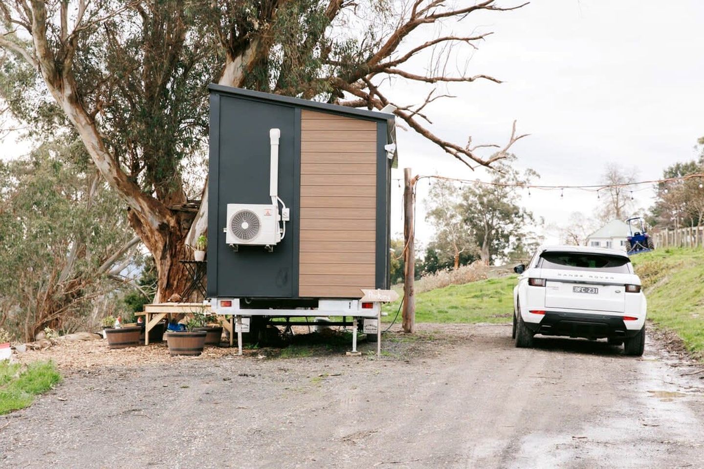 A portable structure with a brown door and a white air conditioning unit, parked on a gravel path with a white vehicle nearby.