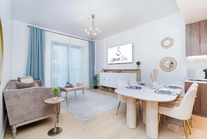A modern living room with a sofa, a dining table set for four, and a kitchen area with a TV mounted on the wall. - bnbme - La Cote 3 - 1BR (Dubai)