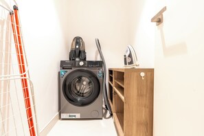 A compact laundry area with a front-loading washing machine, a steam iron, and a drying rack. - bnbme - La Cote 3 - 1BR (Dubai)