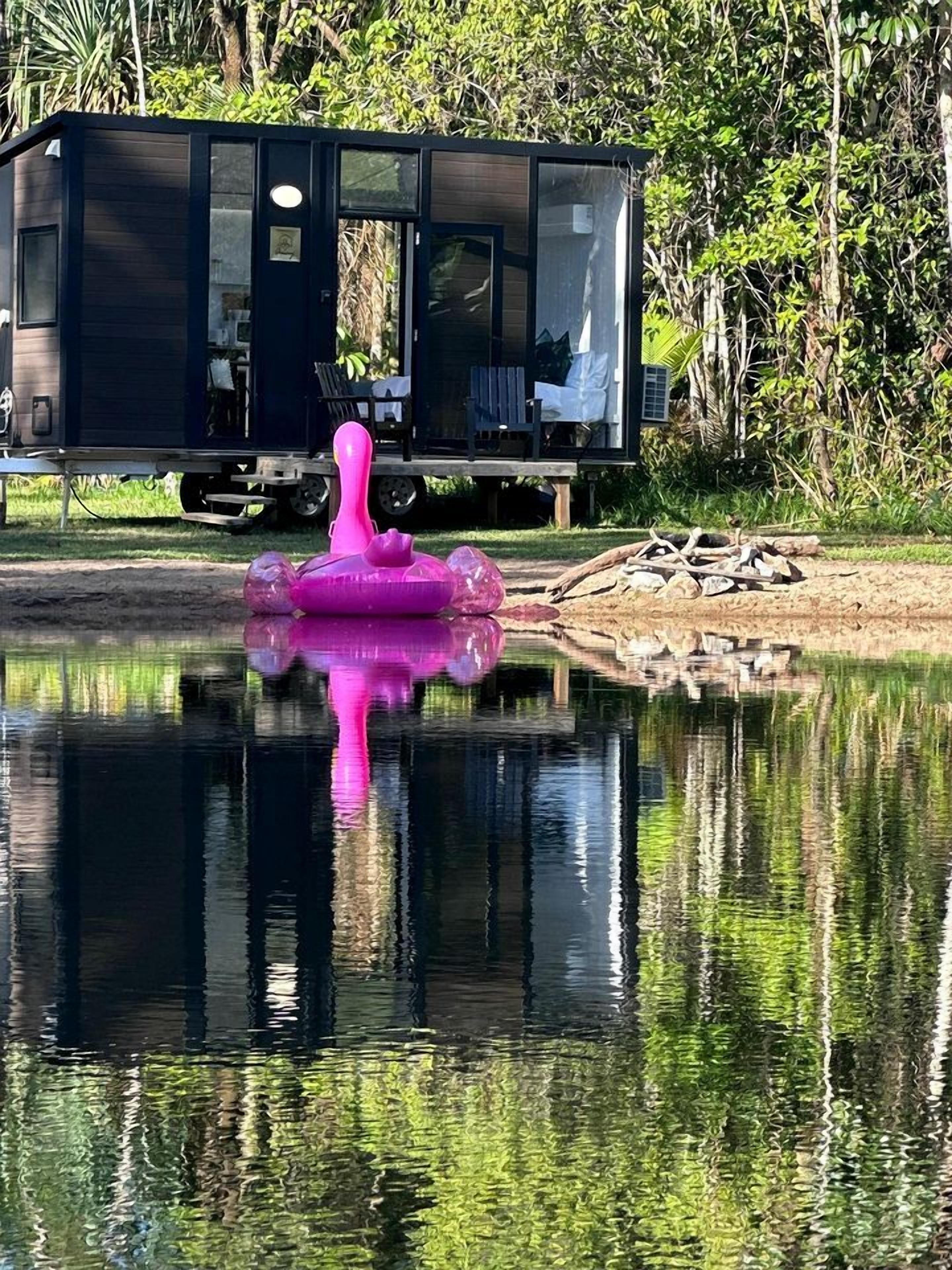 Eine moderne Hütte mit Glastür, gelegen an einem ruhigen Gewässer mit einem rosa Schwimmobjekt.