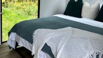 A bedroom with a bed, pillows, a white knitted throw, and a view of greenery outside.