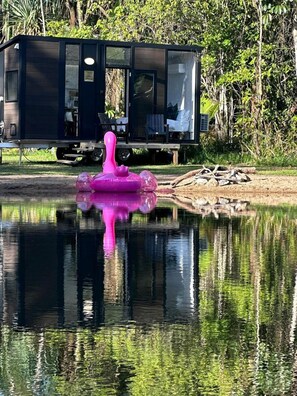 Una cabaña moderna con puerta de cristal, ubicada junto a un cuerpo de agua tranquilo con un objeto inflable rosa.