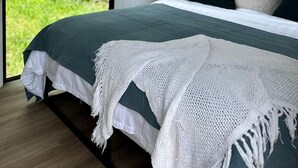 A bedroom with a bed, pillows, a white knitted throw, and a view of greenery outside.