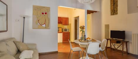 Una sala de estar con sofá, una mesa de comedor con sillas, un cuadro en la pared y una cocina al fondo.