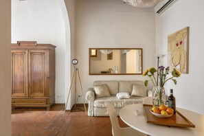 Ein Wohnzimmer mit einer Couch, einem Holzschrank, einem Stativ und einem Tisch mit einer Obstschale und einer Flasche.