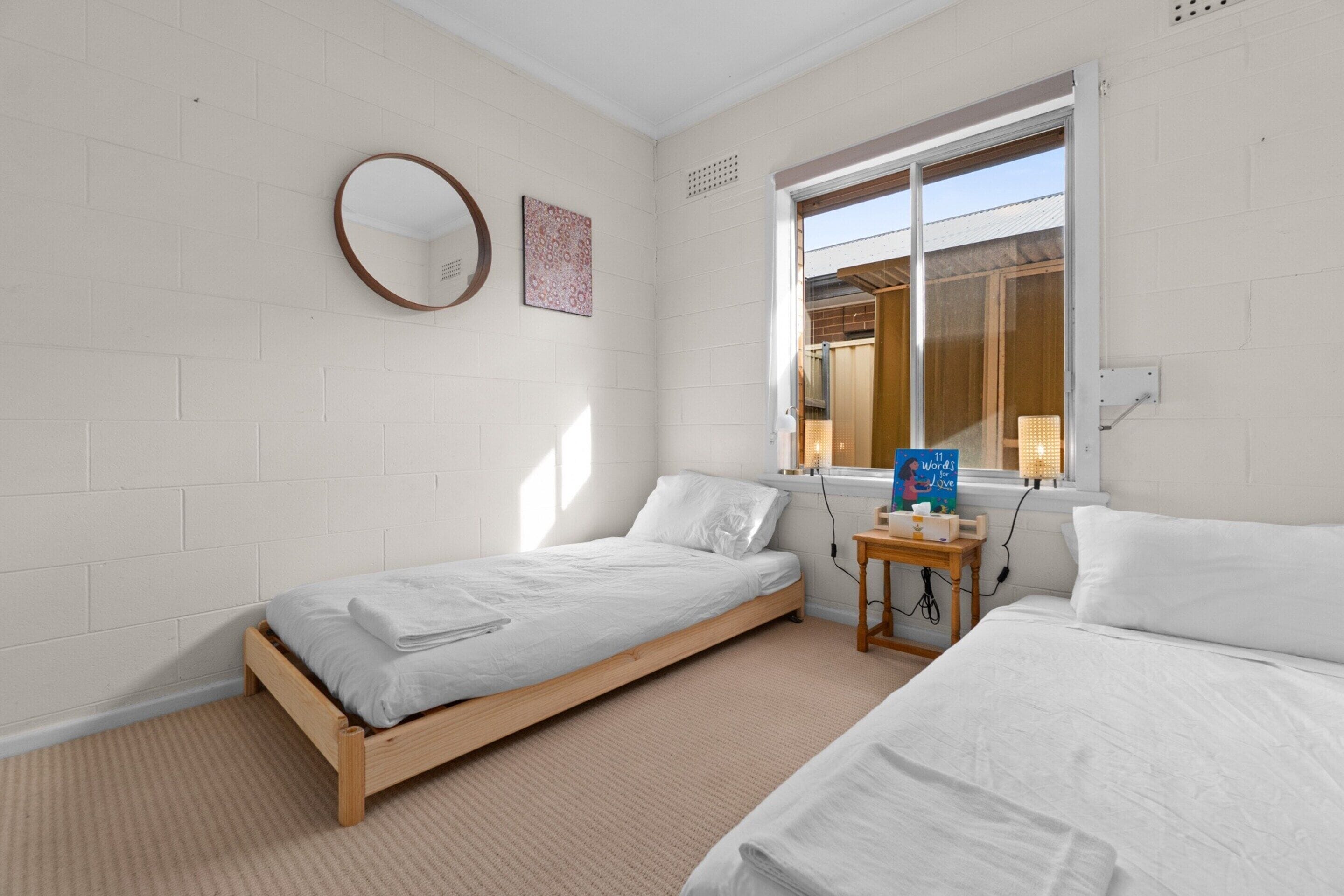 A bedroom with two single beds, a round mirror, a window with a view, and a small table with a book.