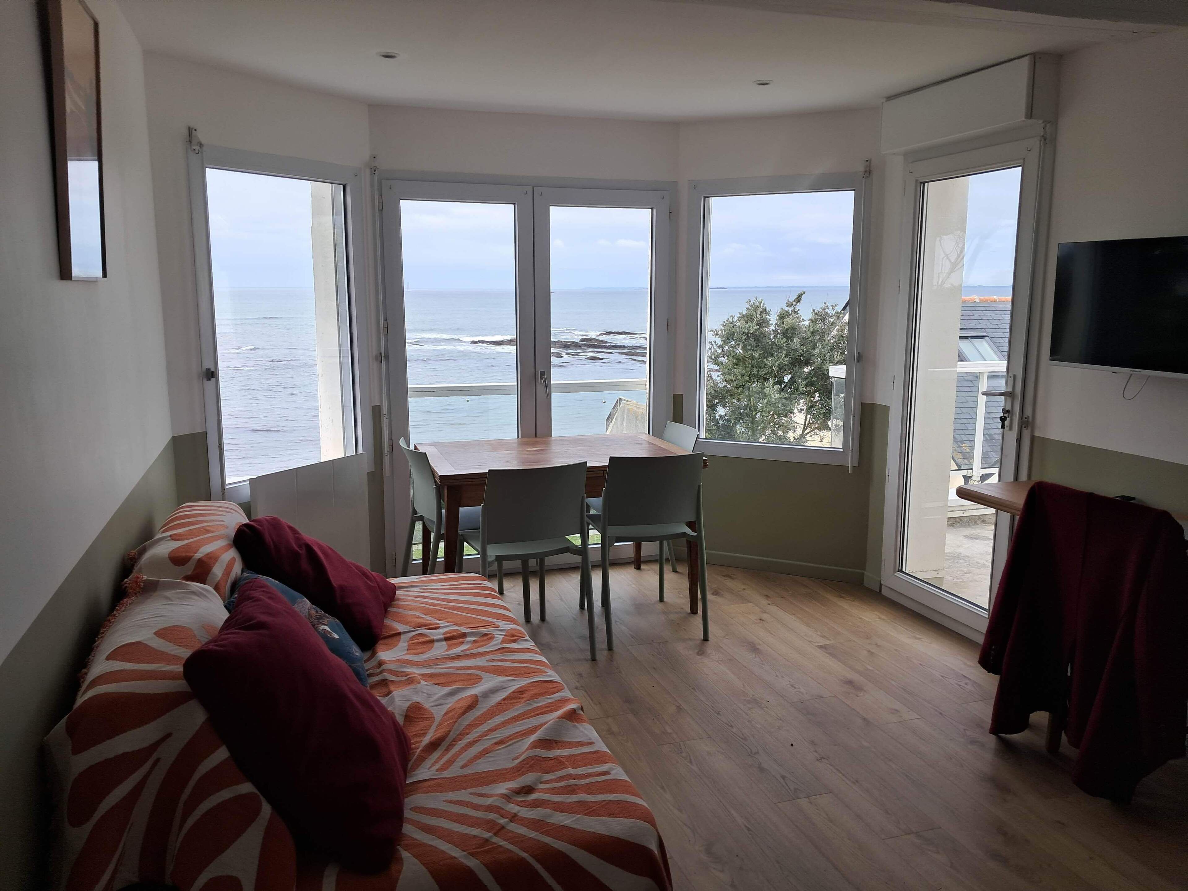 Une pièce comprenant un lit, une table avec des chaises et un téléviseur. De grandes fenêtres offrent une vue sur la mer.
