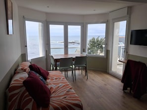 A room with a bed, a table with chairs, and a TV. There are large windows with a view of the sea. - A beautiful 2-room apartment facing the ocean (Piriac-sur-Mer)