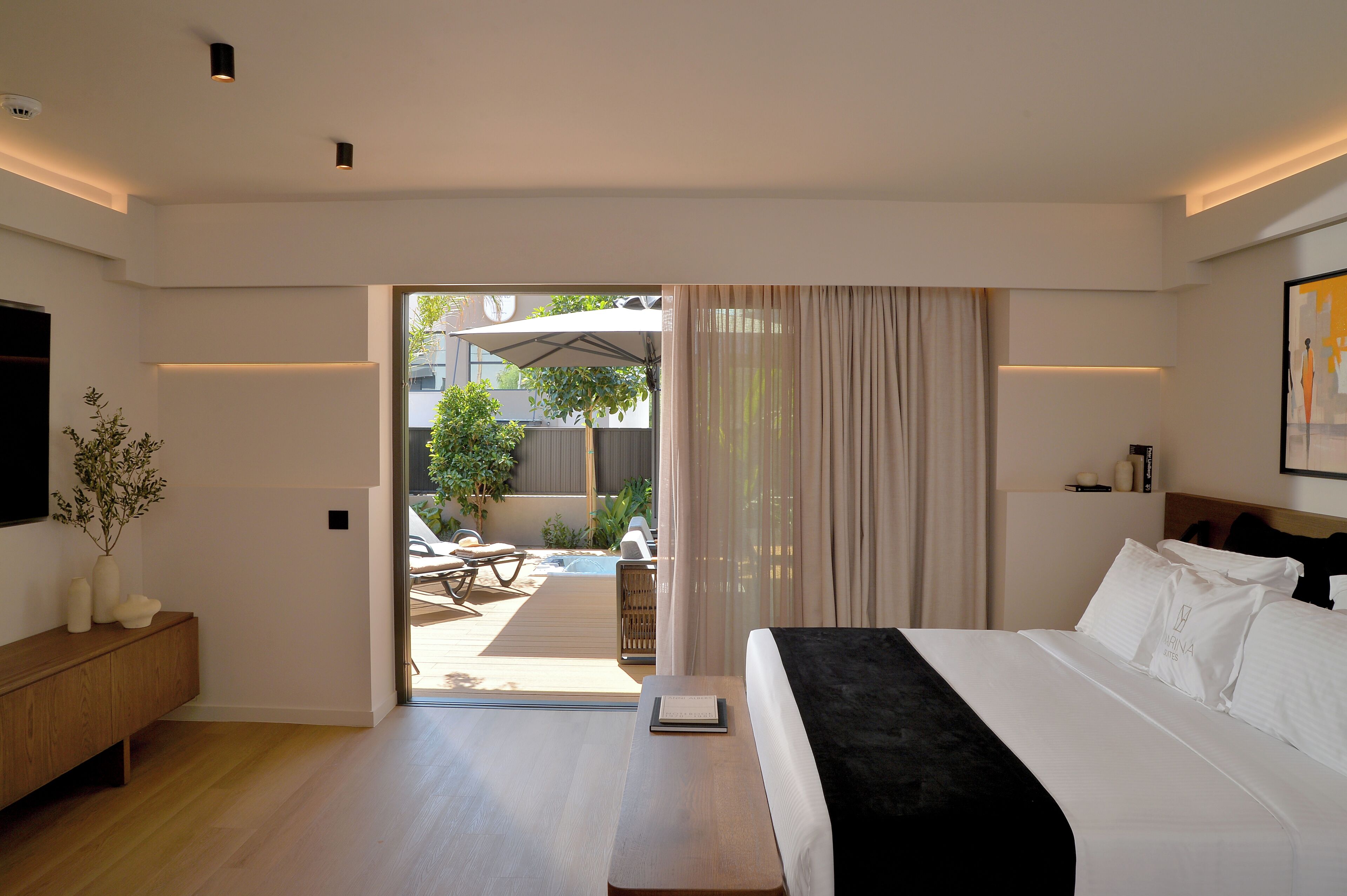 Um quarto moderno com uma cama grande, uma mesa de cabeceira, um criado-mudo e vista para um pátio externo com mesa e cadeiras.