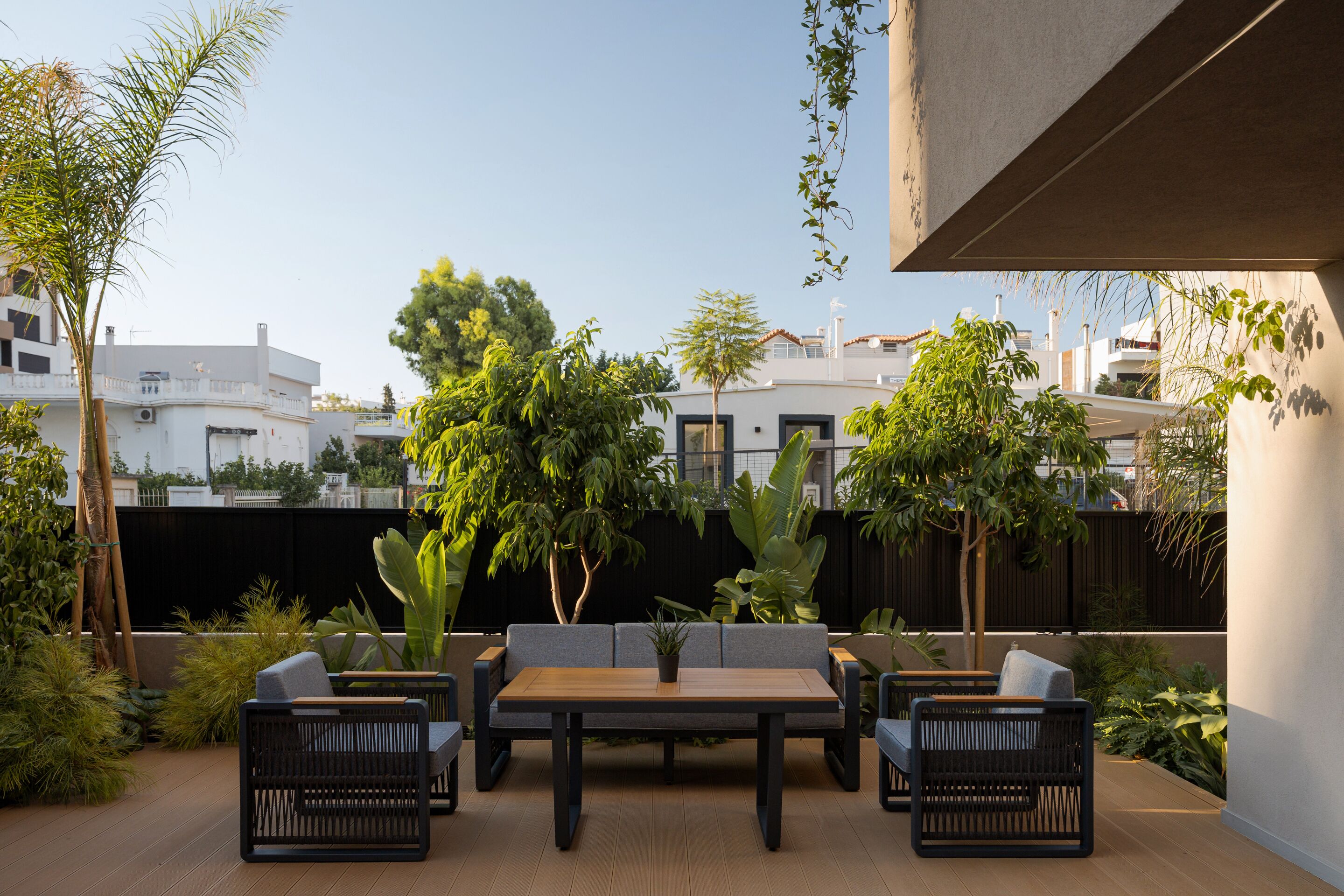 An outdoor patio with a wooden table and two chairs, surrounded by various plants and trees.