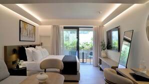A modern hotel room with a bed, a desk, a chair, a small table, and a balcony with a view of greenery.