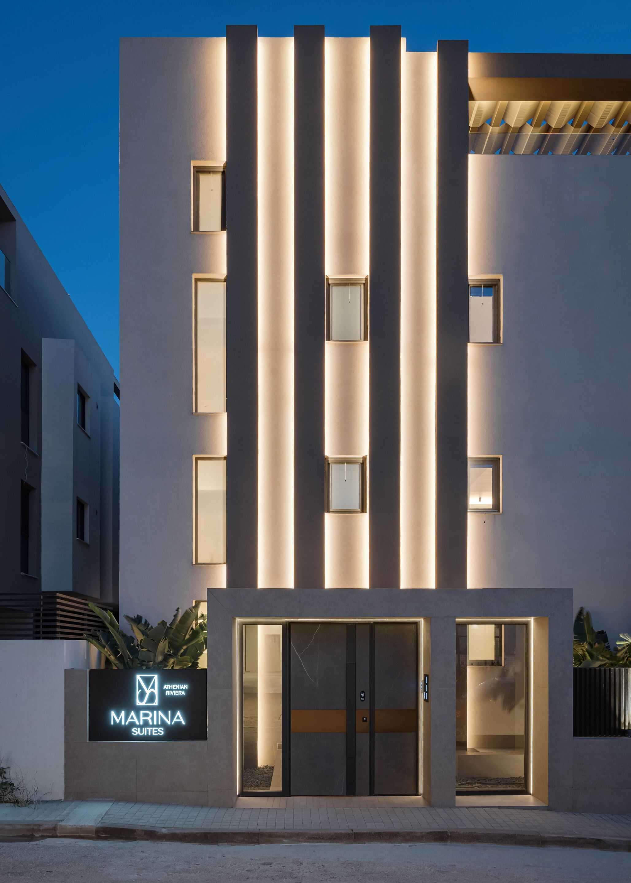 Um edifício moderno com faixas verticais de luz, entrada de vidro e uma placa com 'Marina Suites'.