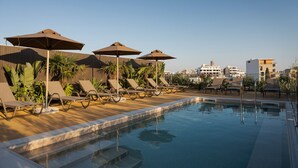 A poolside area with lounge chairs and umbrellas, a clear sky, and buildings in the background.