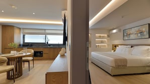 A modern hotel room with a bed, bedside tables, a seating area, a kitchen, and a dining table.