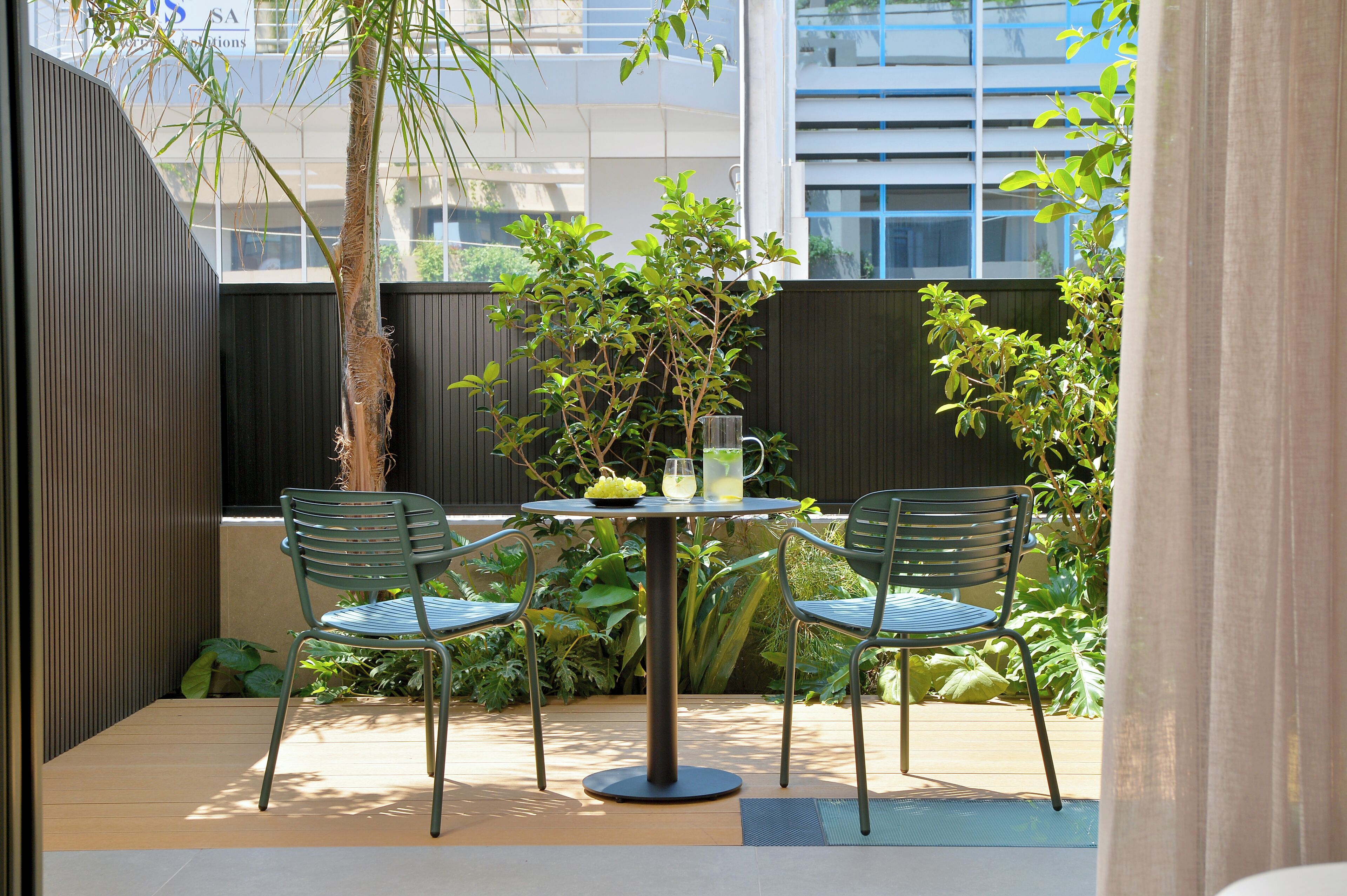 Un patio al aire libre con una mesa redonda, dos sillas, una planta en maceta y una jarra de vidrio.