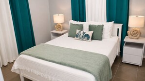 Una cama bien hecha con una manta verde, ropa de cama blanca y almohadones decorativos. Hay dos mesitas de noche con lámparas y conchas decorativas. La habitación tiene cortinas color turquesa y paredes blancas.