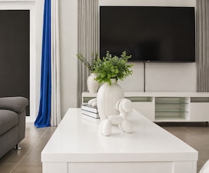 A modern living room with a white coffee table, a white vase with green plants, a white dog sculpture, a grey sofa, and a flat-screen TV mounted on the wall. - Chic Blue Oasis Modern Curaçao Willemstad Retreat (Willemstad)